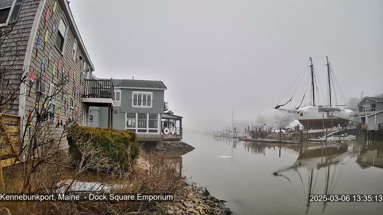 A foggy waterfront scene shows a gray building with colorful decorations, another gray building with a "LOVE" sign, and a large sailboat docked, all reflected in calm water.