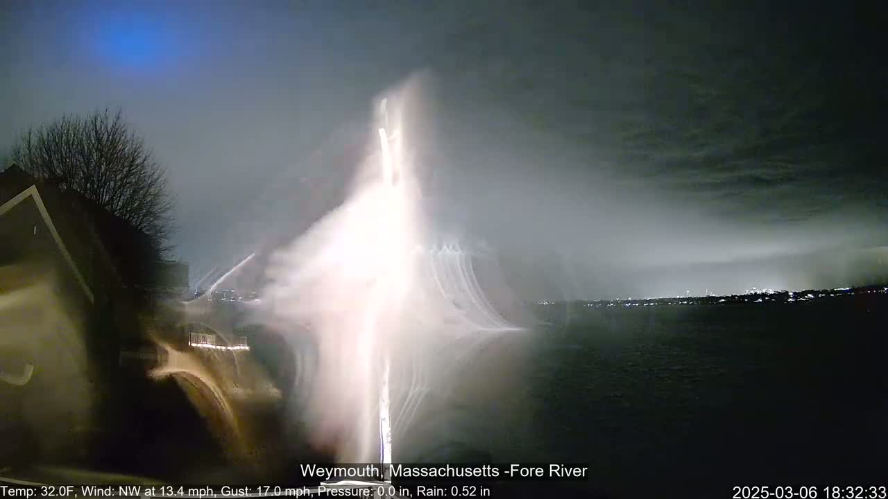 A nighttime view of a bright, blurry light source over a dark body of water under a cloudy sky with light rain.