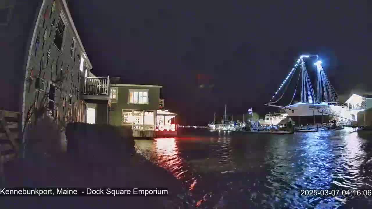 A large, illuminated ship is docked at a waterfront at night, next to smaller boats and buildings with lights on.