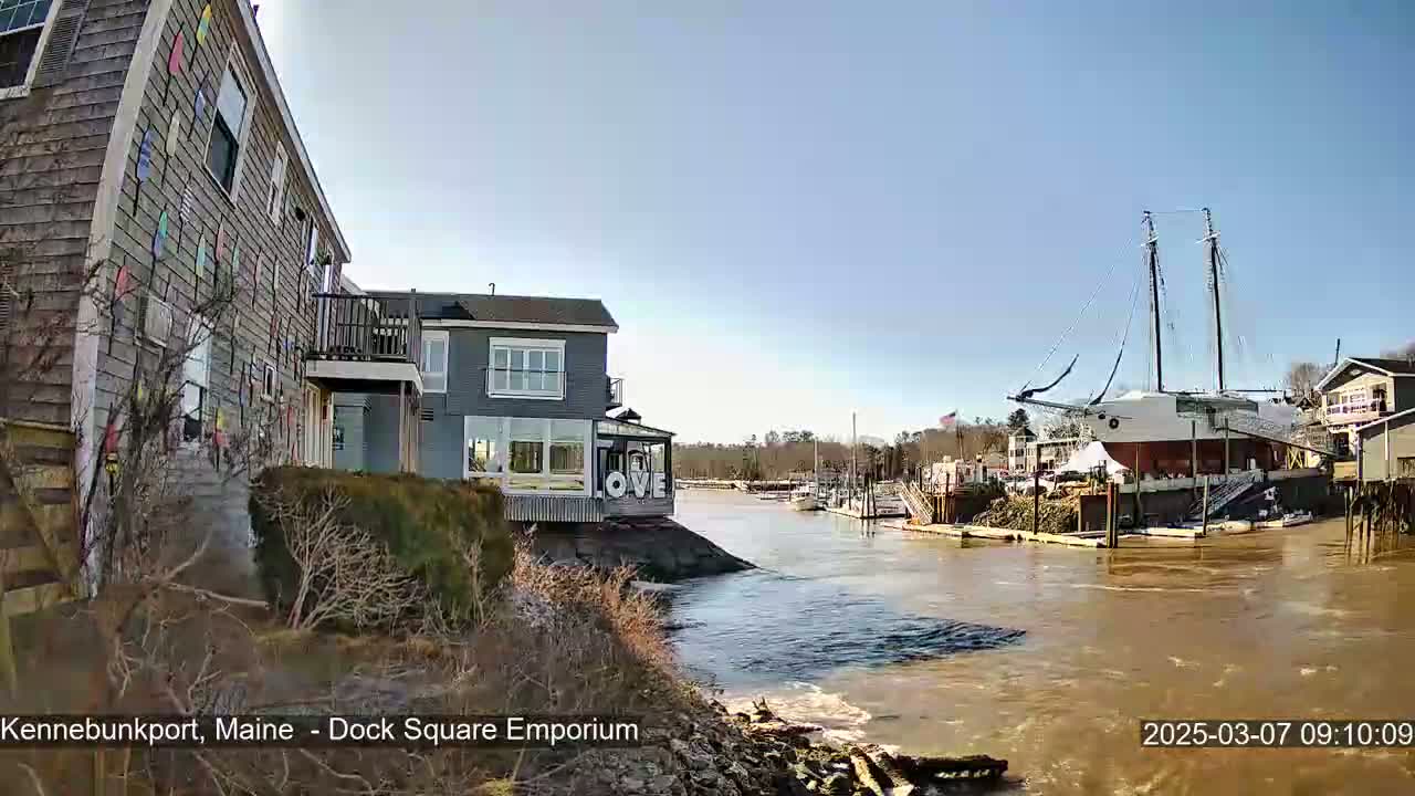 A waterfront view on a sunny day shows a gray building with a "LOVE" sign, other buildings, a large docked sailboat, and a muddy waterway.