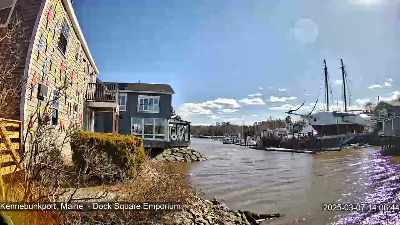A waterfront scene on a sunny day shows a building decorated with colorful nautical flags next to a calm river with a large sailing ship docked nearby.