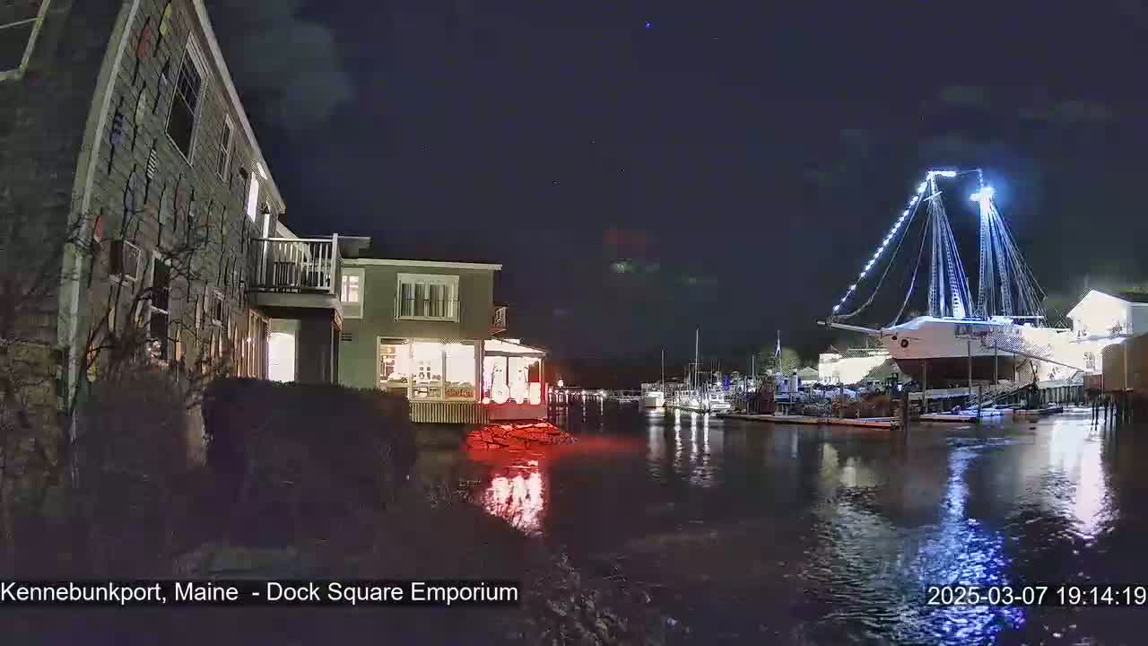 A large sailboat, illuminated with white lights, is moored in a calm harbor at night next to a waterfront building.