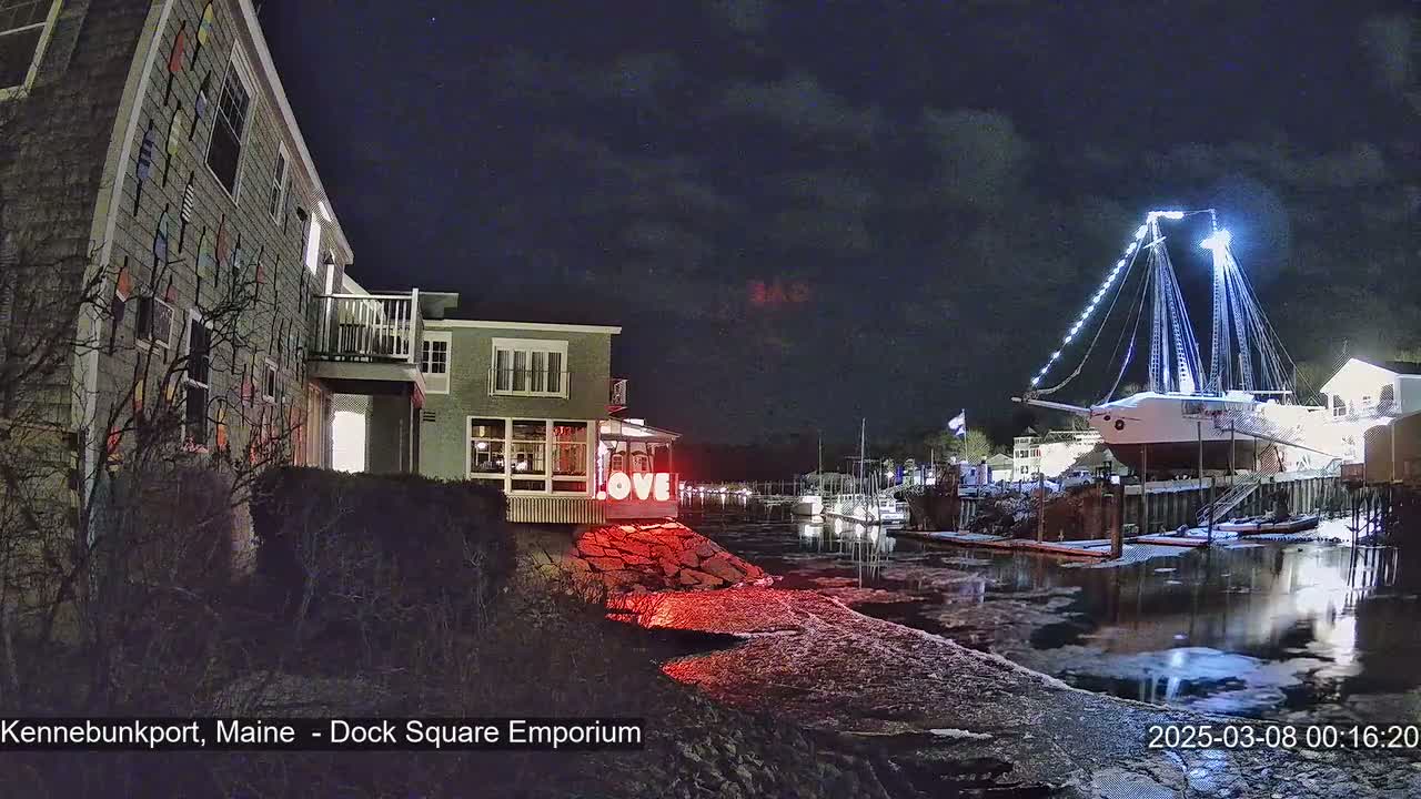 A nighttime waterfront scene shows a building decorated with colorful buoys, a lit-up sailboat docked nearby, and a calm waterway under a partly cloudy sky.