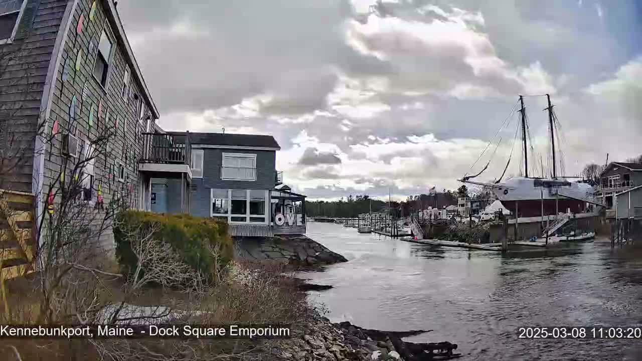 A waterfront scene under a cloudy sky shows a canal with docked boats, including a large sailing ship, and buildings along the shore, one of which has colorful decorations on its exterior.