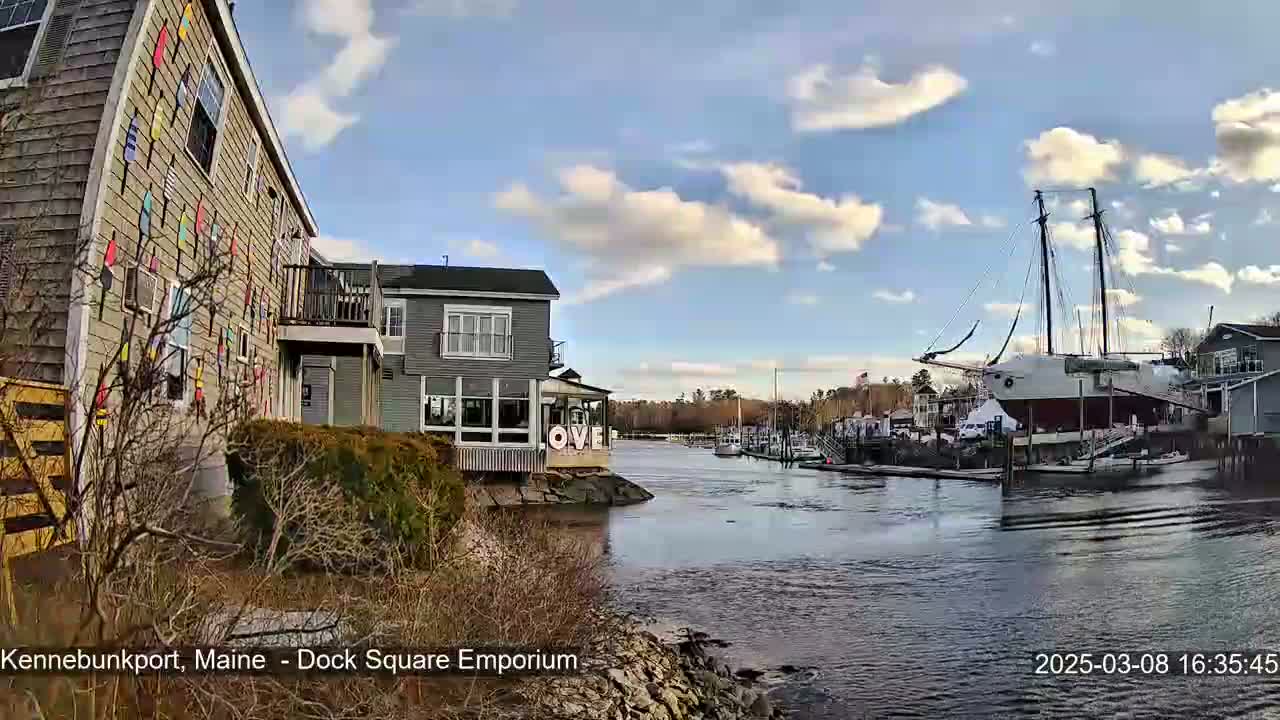 A partly cloudy blue sky overlooks a waterfront scene with buildings, docked boats, and a large sailing ship, all situated along a calm body of water.