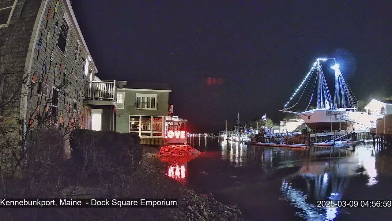 A large, illuminated sailboat is docked at night next to a building with colorful exterior decorations, all reflected in calm water.