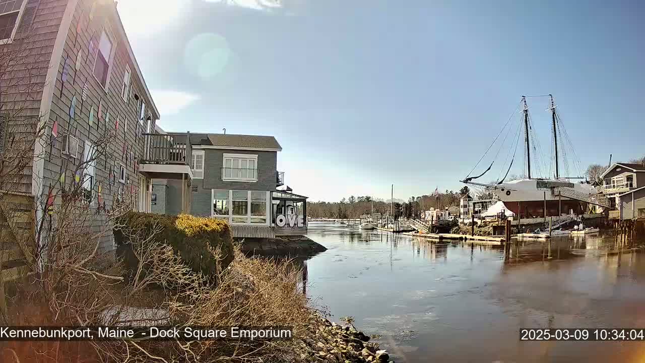 A waterfront scene on a sunny day shows a gray building decorated with nautical flags, another building with large windows, and a large sailboat docked in the calm water of a harbor.