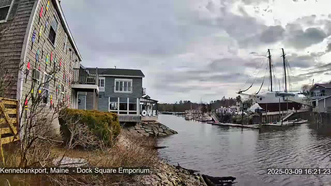 A waterfront view on an overcast day shows a building with colorful artwork on its exterior, other buildings along a calm waterway, and a large sailboat docked nearby.