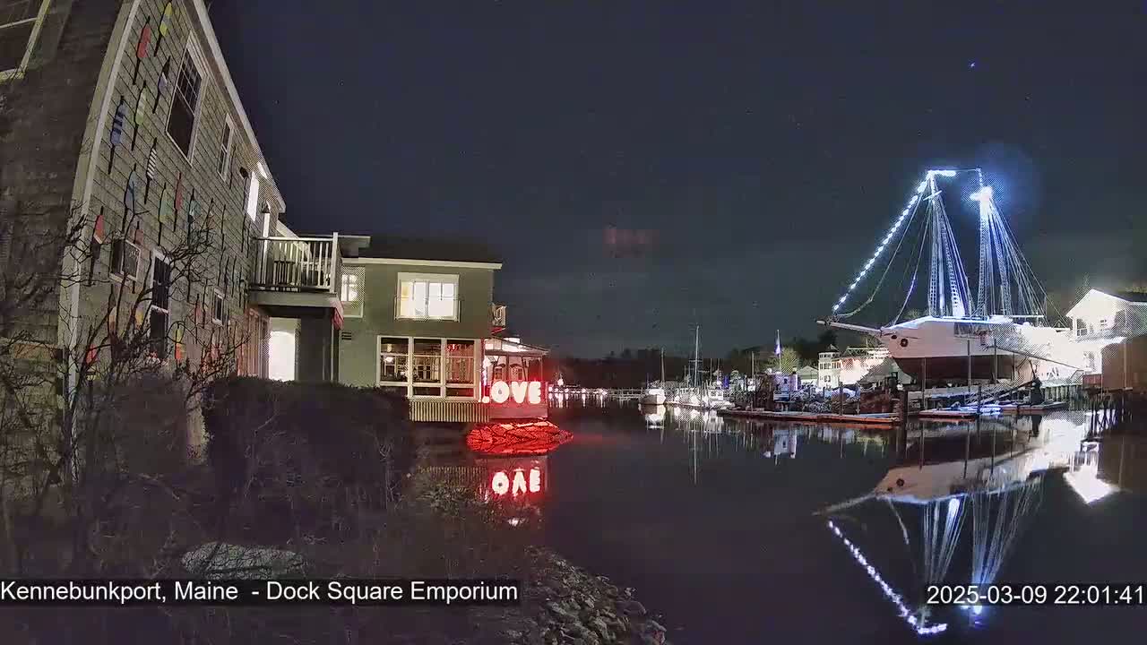 A nighttime view of a harbor with a large, illuminated ship, buildings, and a "LOVE" sign reflected in the calm water under a clear, starry sky.