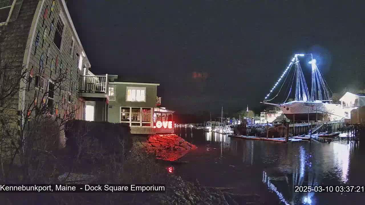 A nighttime waterfront scene shows a building decorated with nautical flags, a large ship illuminated with lights, and a "LOVE" sign, all reflected in dark water.