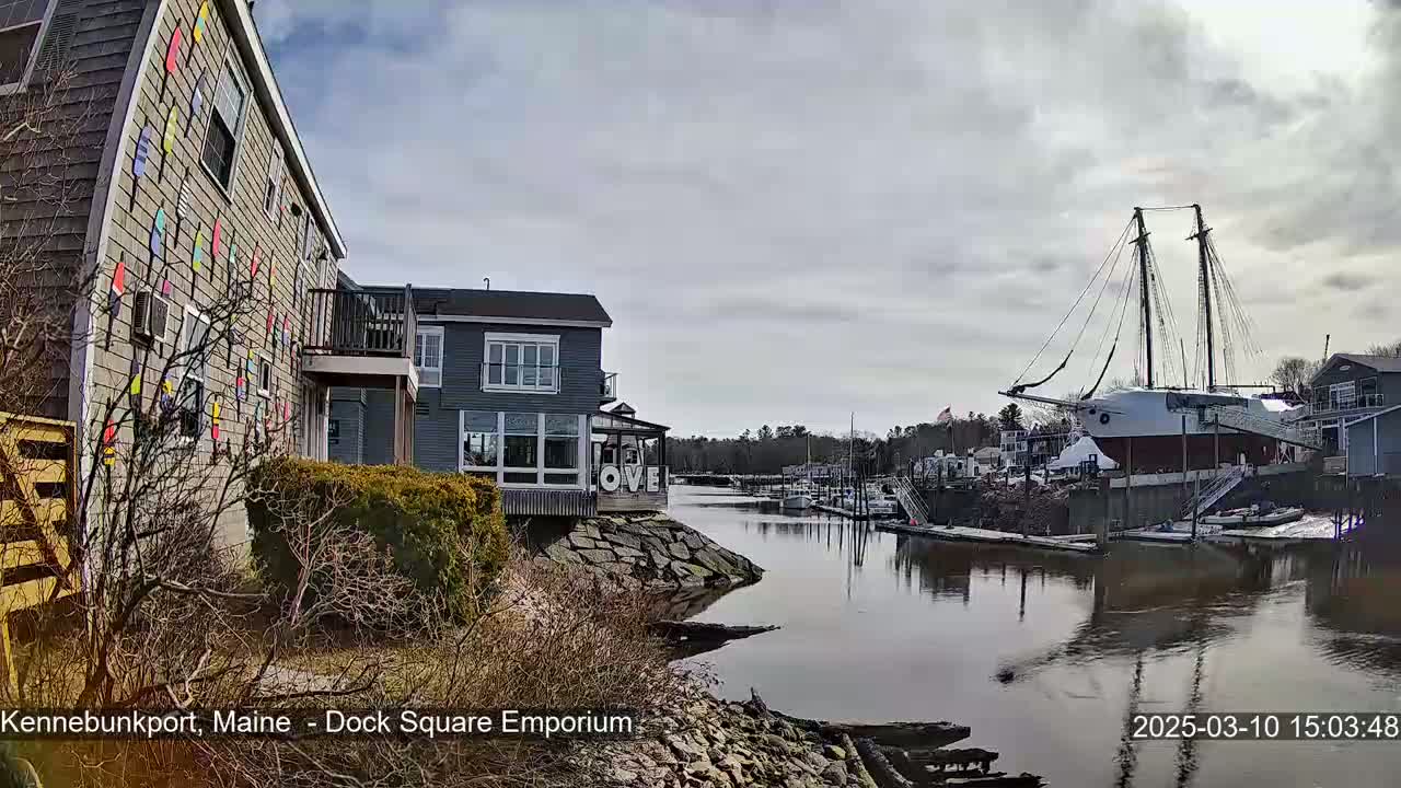 A calm river flows past buildings, one with colorful paddle-like decorations on its exterior, toward a large docked sailing ship under a cloudy sky.