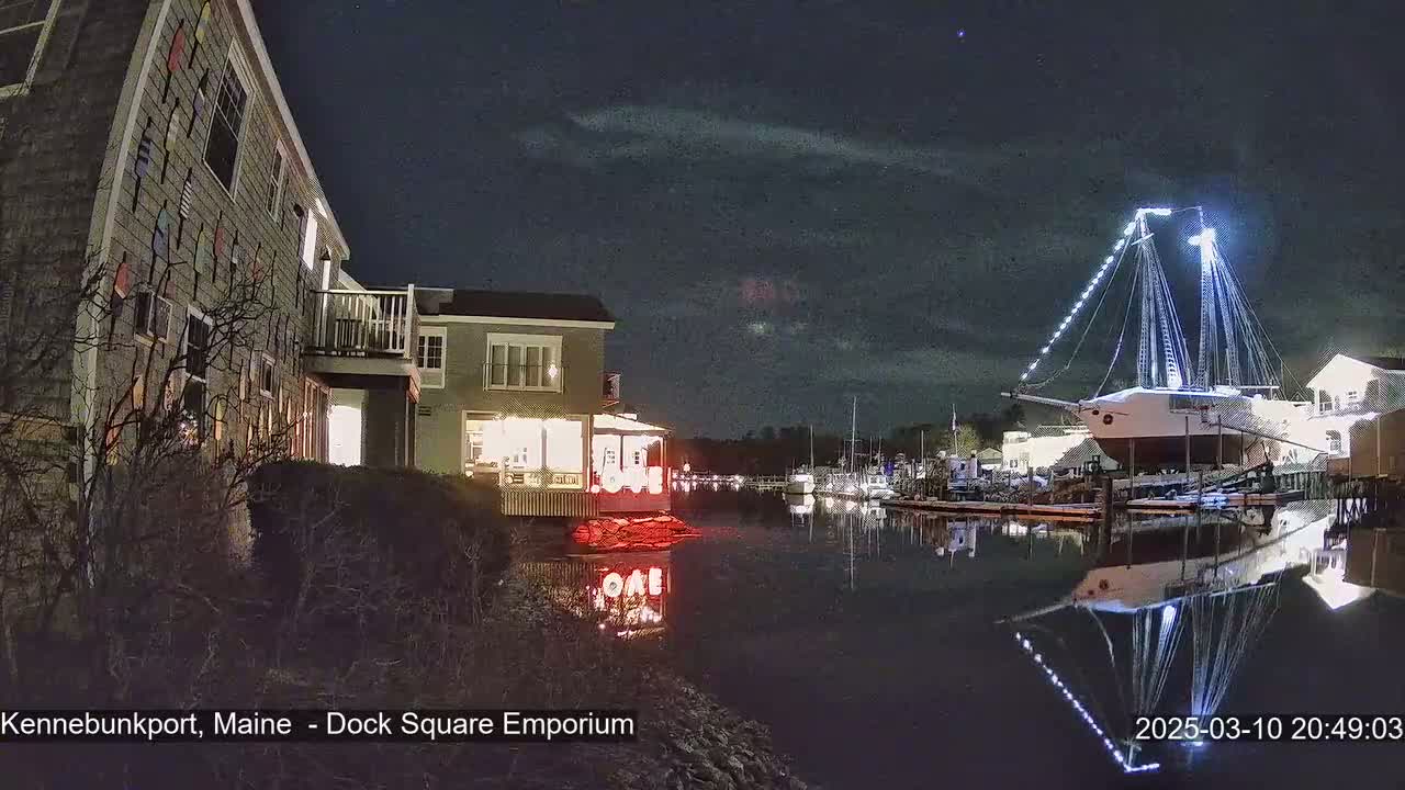 A nighttime scene shows a waterfront with a large sailboat decorated with lights, reflecting in calm water, next to buildings illuminated along a dock.