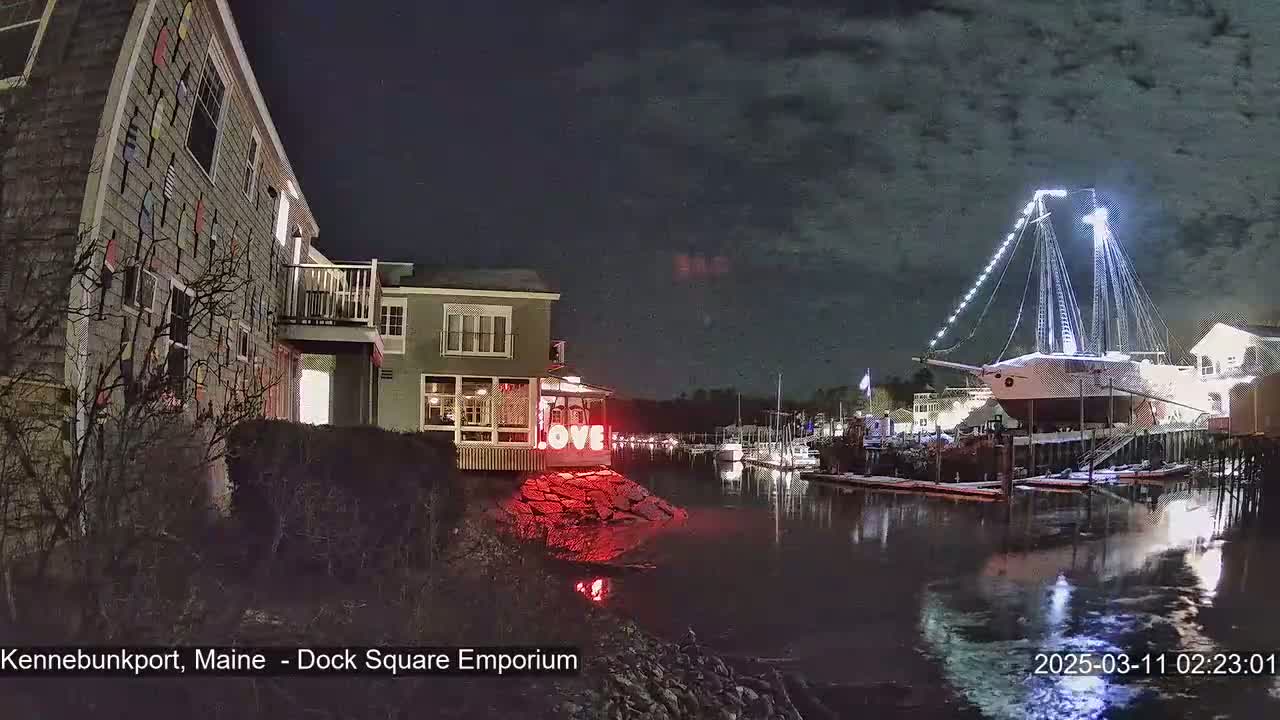 A large illuminated sailboat is docked at night next to a building with lights and the word "LOVE" displayed, all reflected in calm water under a cloudy sky.