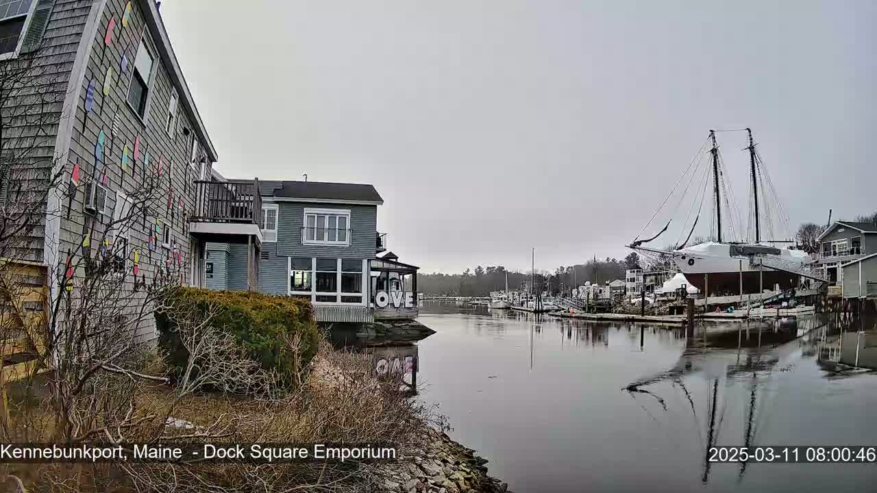 A gray, overcast day shows a waterfront scene with buildings, a large sailboat docked, and calm water reflecting the buildings and boats.