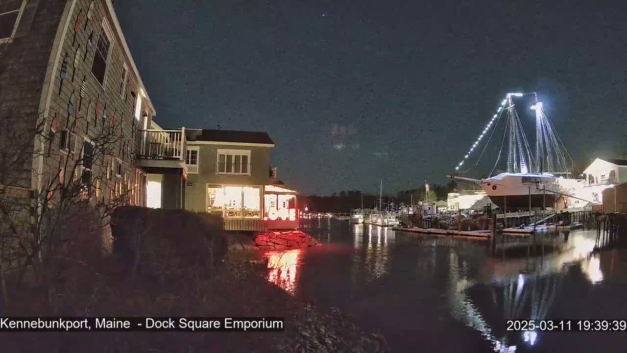A large illuminated ship is docked at a waterfront at night, next to buildings with lights on.