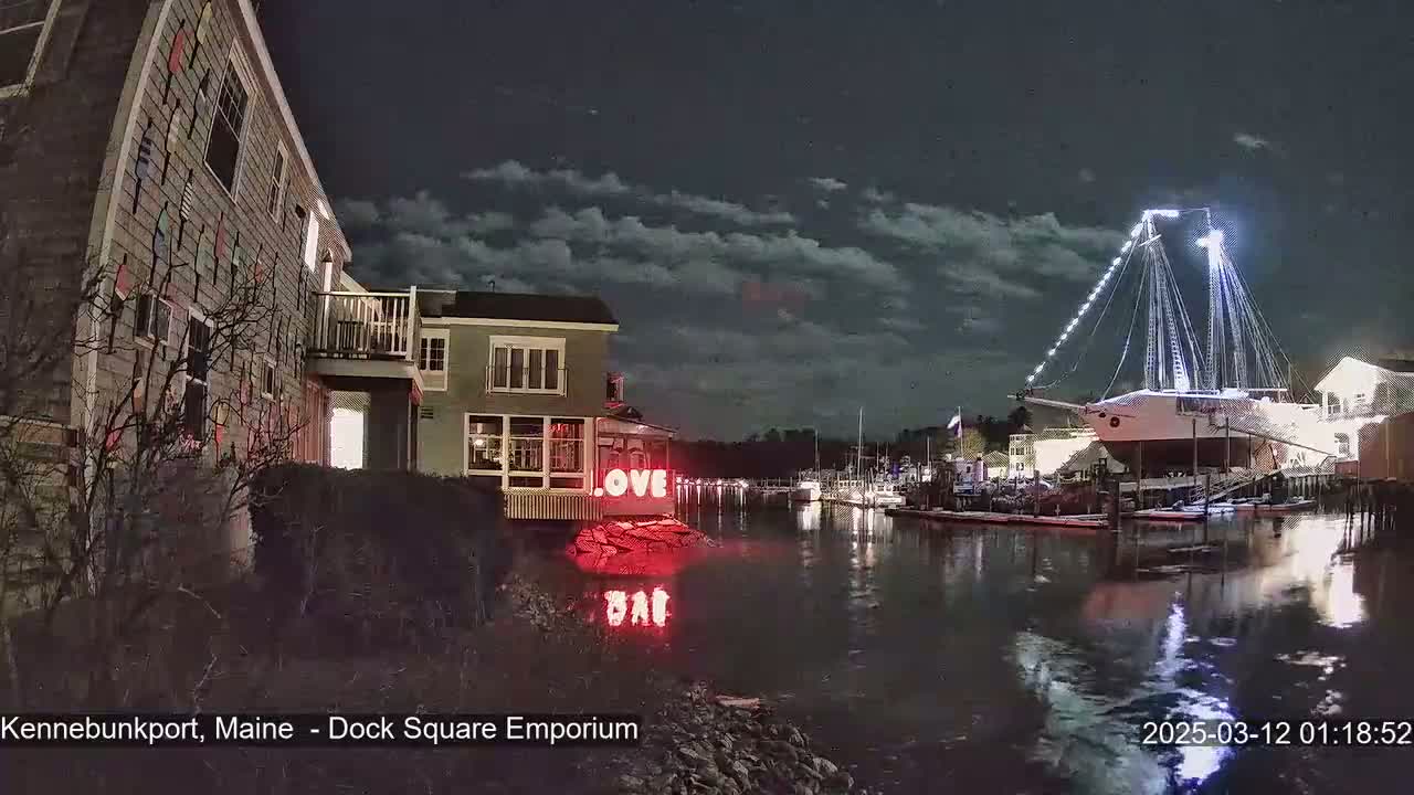 A nighttime view of a harbor with a large sailboat docked, lit buildings along the shore, and a "LOVE" sign on a building near the water under a partly cloudy sky.