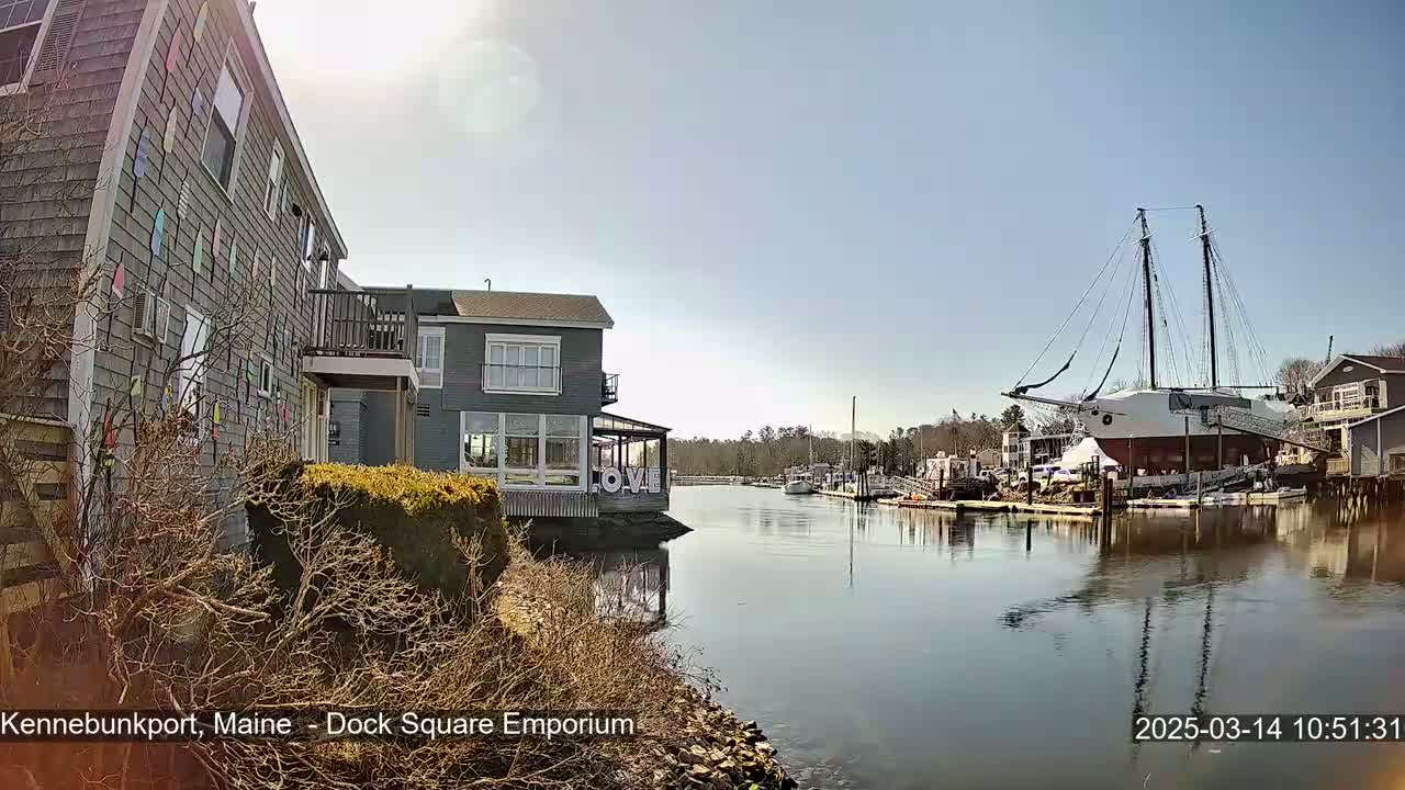 A calm waterway reflects a large sailing ship docked alongside buildings, under a sunny, clear sky.