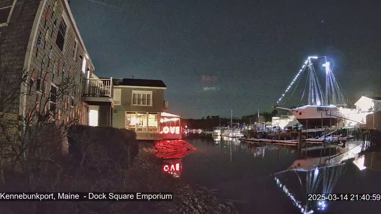 A nighttime harbor scene shows a large sailing ship docked near buildings decorated with lights, under a dark, clear sky.