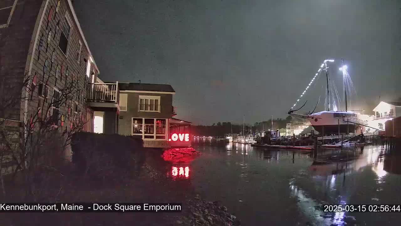 A snow-covered sailboat is docked at night near a building with a red "LOVE" sign, illuminated against a dark, snowy sky.
