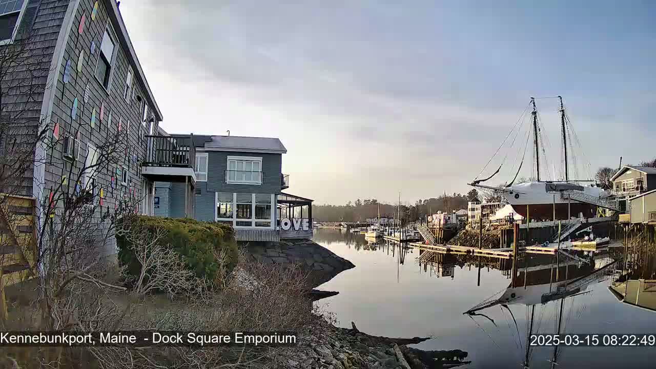 A calm harbor scene under a clear sky shows buildings along the water's edge, a large sailboat docked, and smaller boats moored nearby.