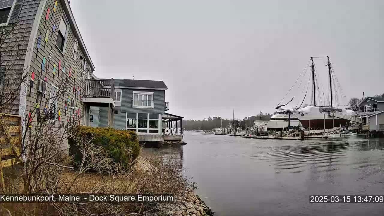 A gray overcast day shows a waterway with docked boats, including a large sailboat, and buildings along the shore, one of which has colorful paddle-shaped decorations on its exterior.