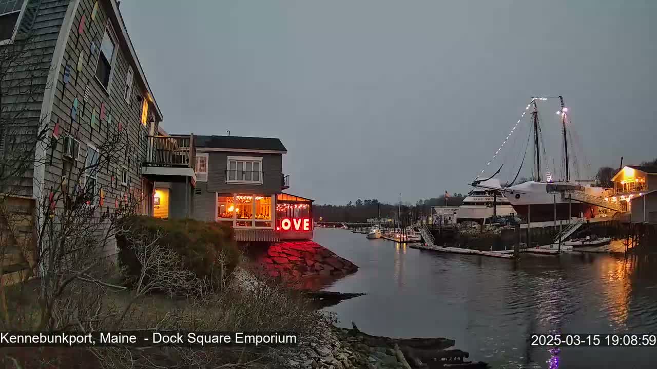 A waterfront scene at dusk shows a building with illuminated "LOVE" signage, other buildings, boats docked at a pier, and a large sailboat under a cloudy sky.