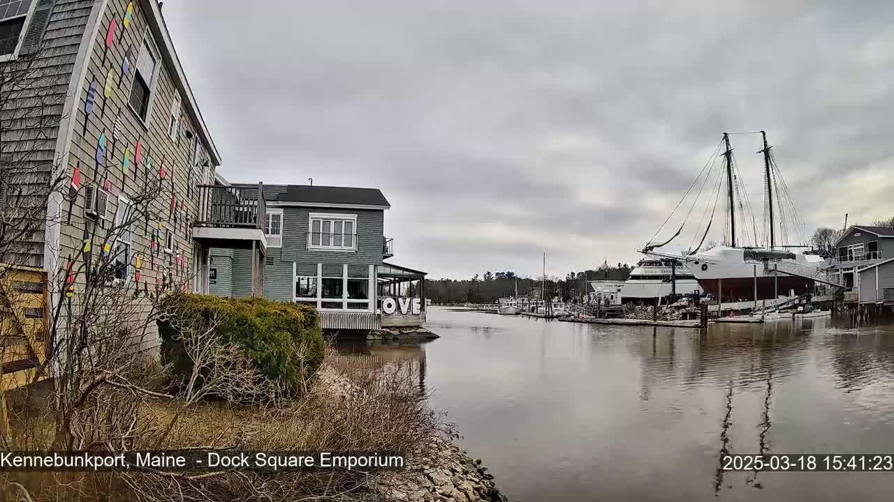 A calm waterway reflects a cloudy sky, with buildings and a large sailboat visible along the shore.