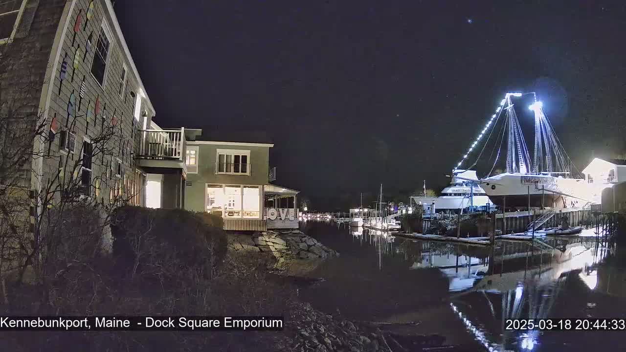 A large sailboat is docked at night next to a waterfront building, with its reflection visible in the calm water.