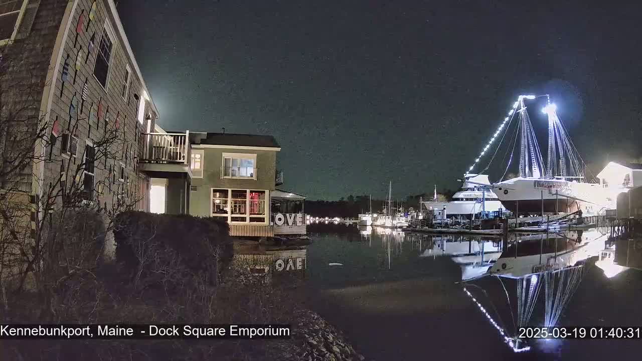 A nighttime view of a harbor, showing a building with nautical decorations, another building with lit windows, and a large sailboat illuminated with lights, all reflected in calm water under a clear, starry sky.