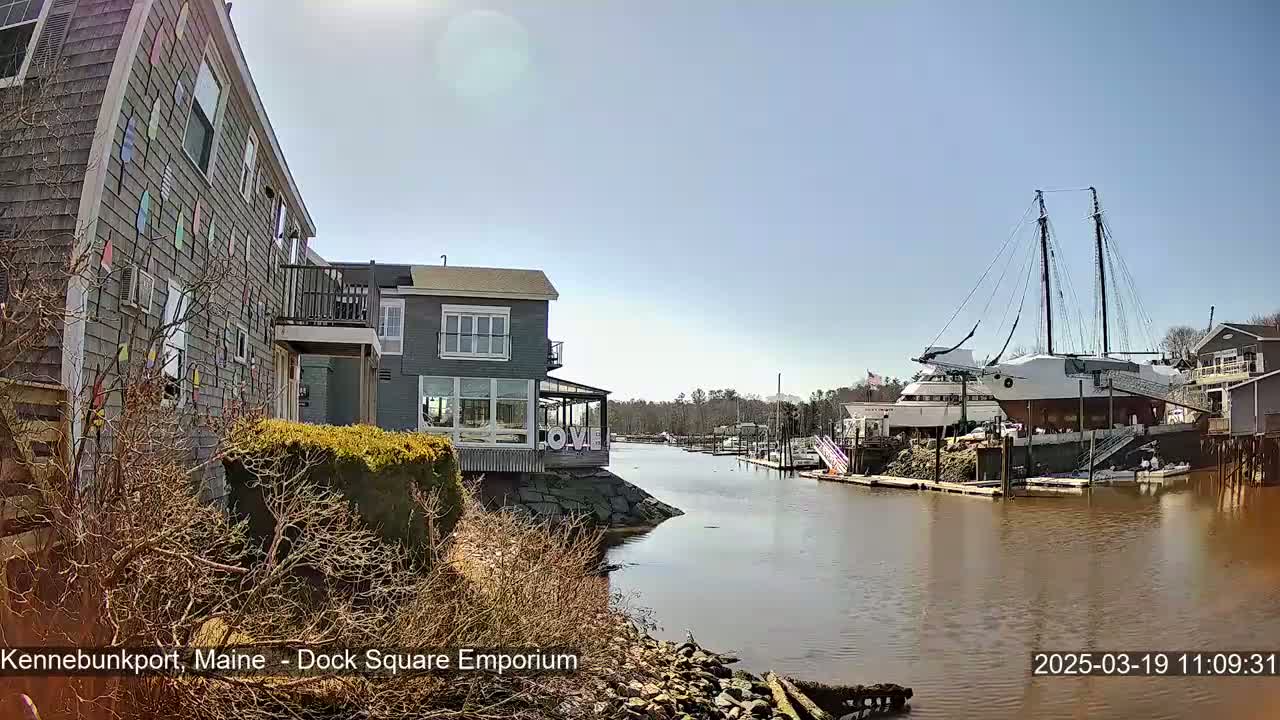 A waterfront view on a sunny day shows a gray building with colorful decorations on its exterior, another gray building with large windows, and a large sailboat docked at a nearby pier.