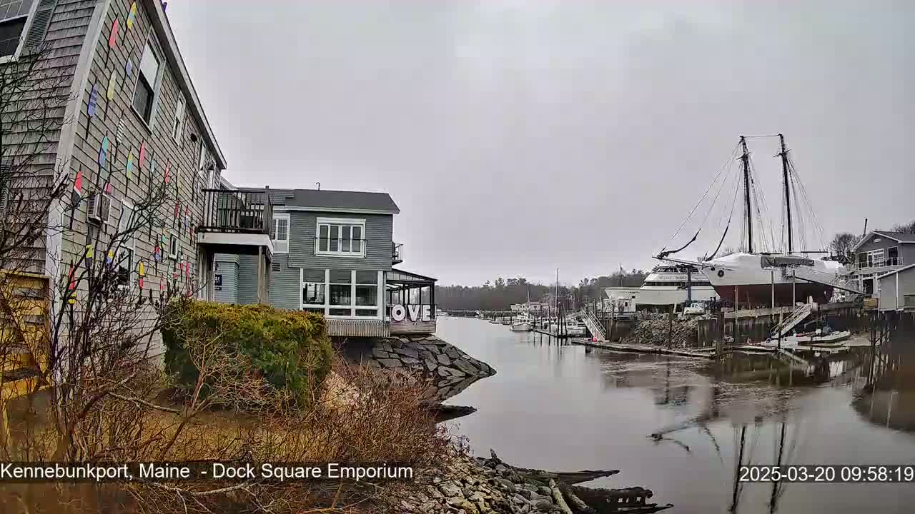 A gray, overcast day reveals a waterfront scene with buildings, a large sailboat docked, and a calm river.