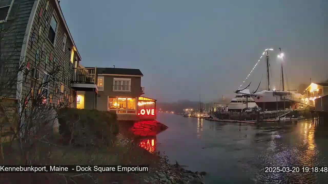 A waterfront scene at twilight shows a building with illuminated windows and a "LOVE" sign, next to a harbor where several boats are docked under a hazy sky.