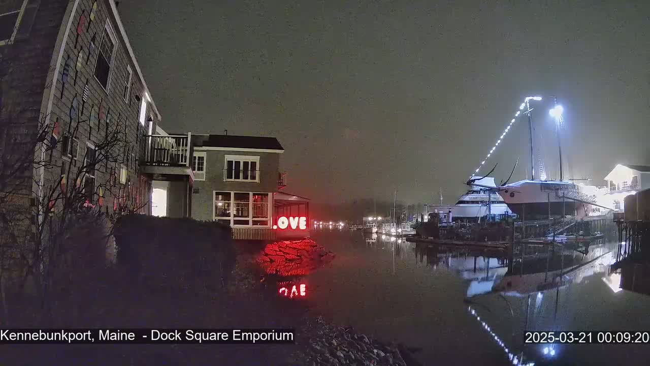A nighttime scene shows buildings along a waterfront canal under a lightly overcast sky, with a large sailboat docked and illuminated, and a red "LOVE" sign near a building.