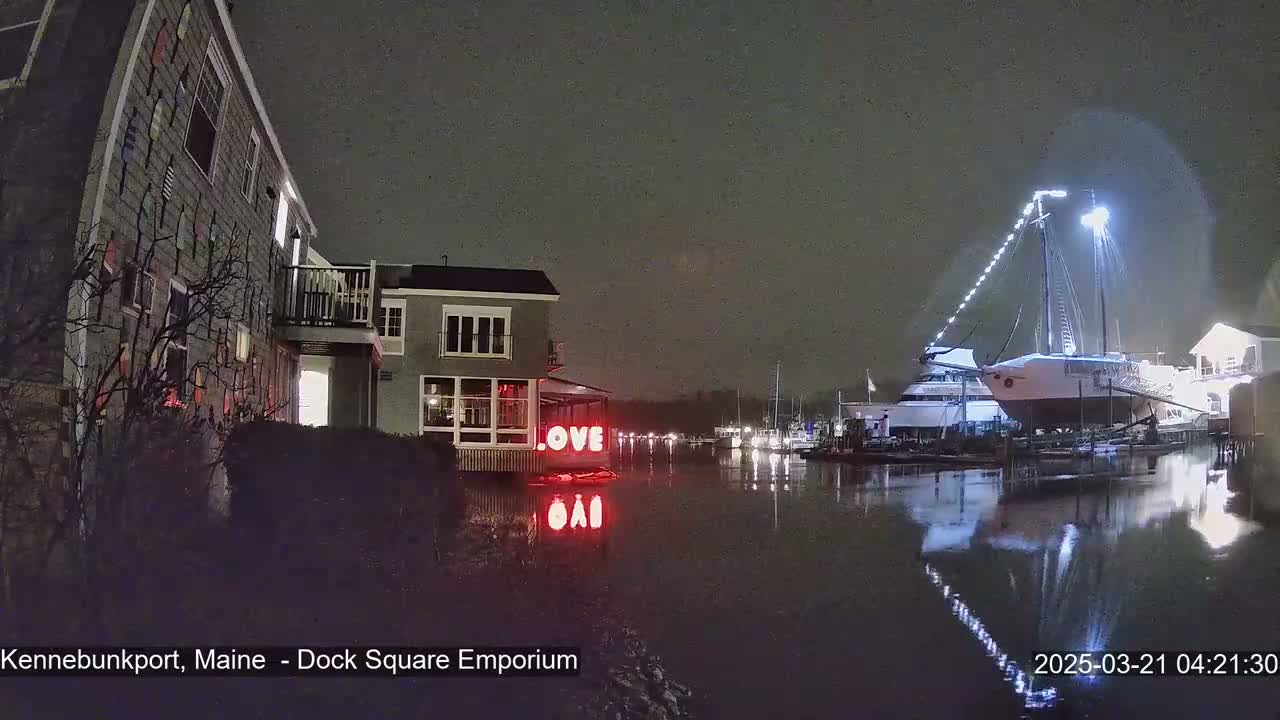 A nighttime waterfront scene shows a building with the word "LOVE" illuminated on its exterior, reflecting in the calm water alongside a docked sailboat, under a dark sky.