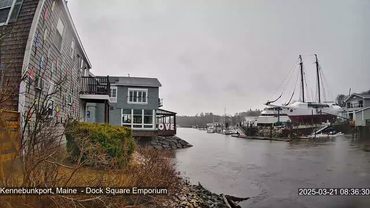 A gray day shows a waterfront scene with buildings, a large docked sailing ship, and several smaller boats, all under an overcast sky.
