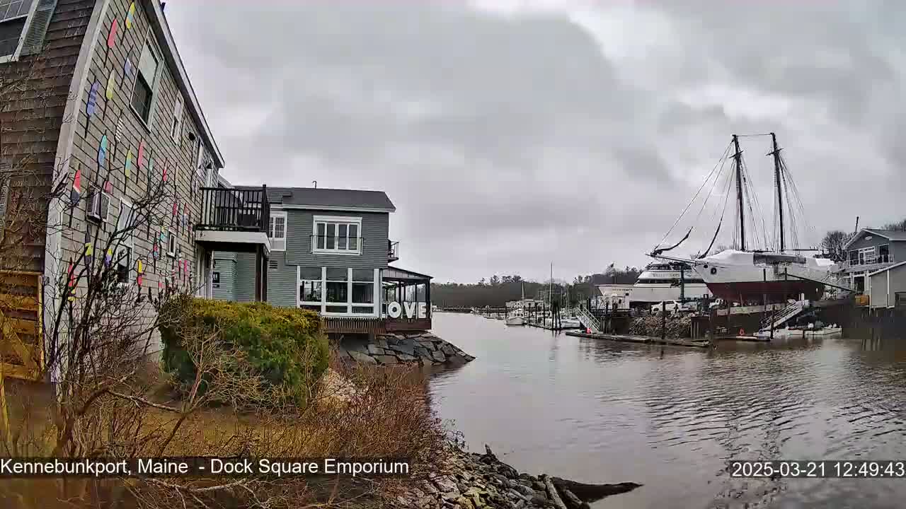 A gray, overcast day shows a waterfront view of buildings, a large sailing ship docked, and a calm waterway.