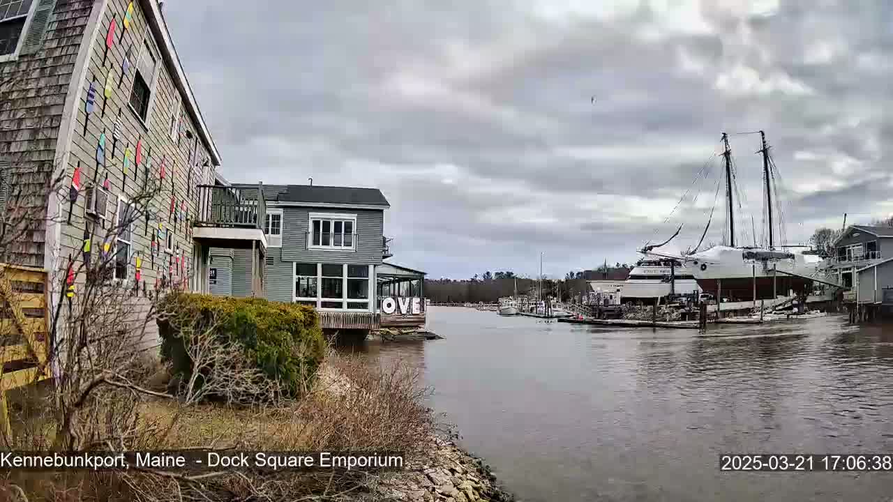 A gray, overcast day reveals a waterfront scene with a building decorated with colorful paddles, another building with large windows, and a large sailboat docked on a calm waterway.