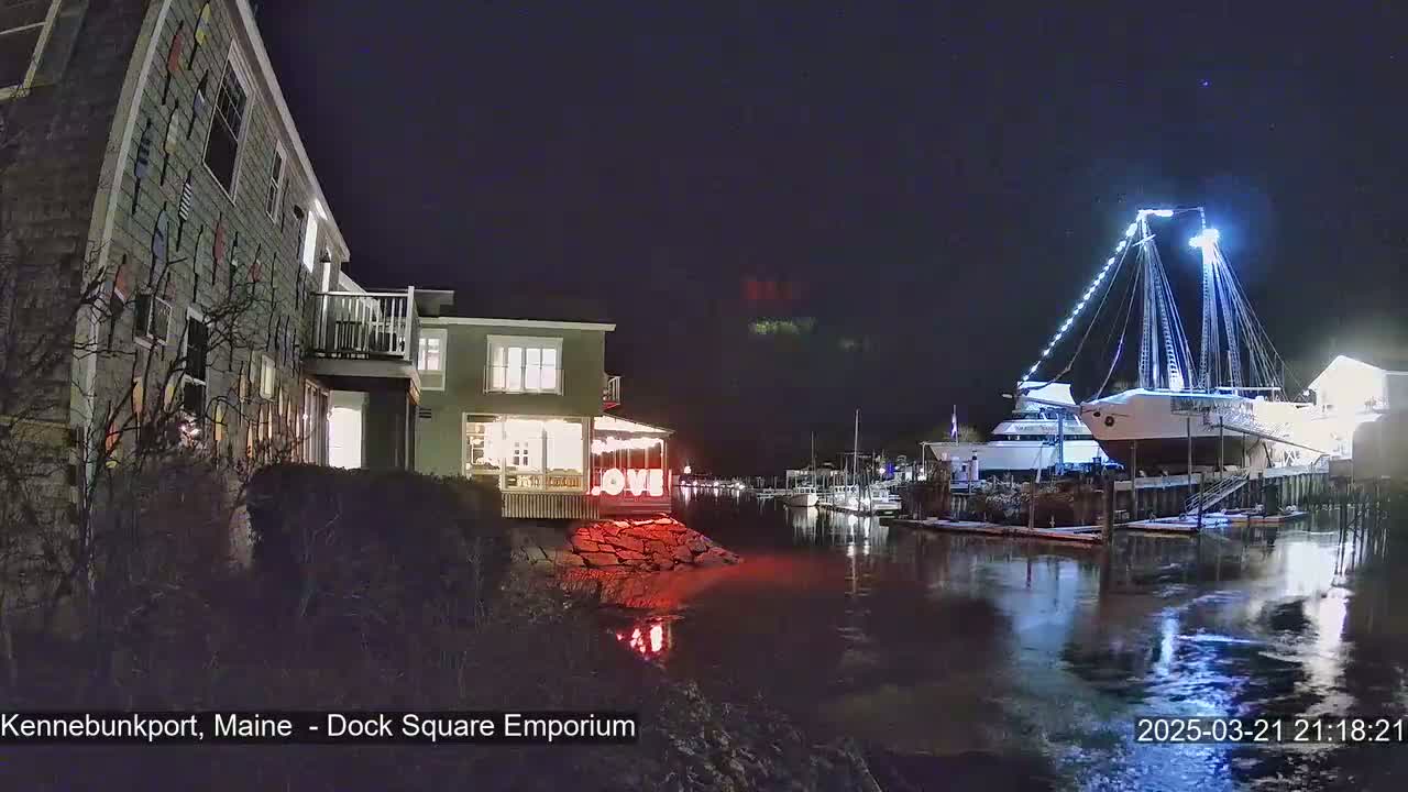 A nighttime harbor scene shows a building decorated with nautical flags, a large illuminated sailboat docked, and calm water reflecting lights.