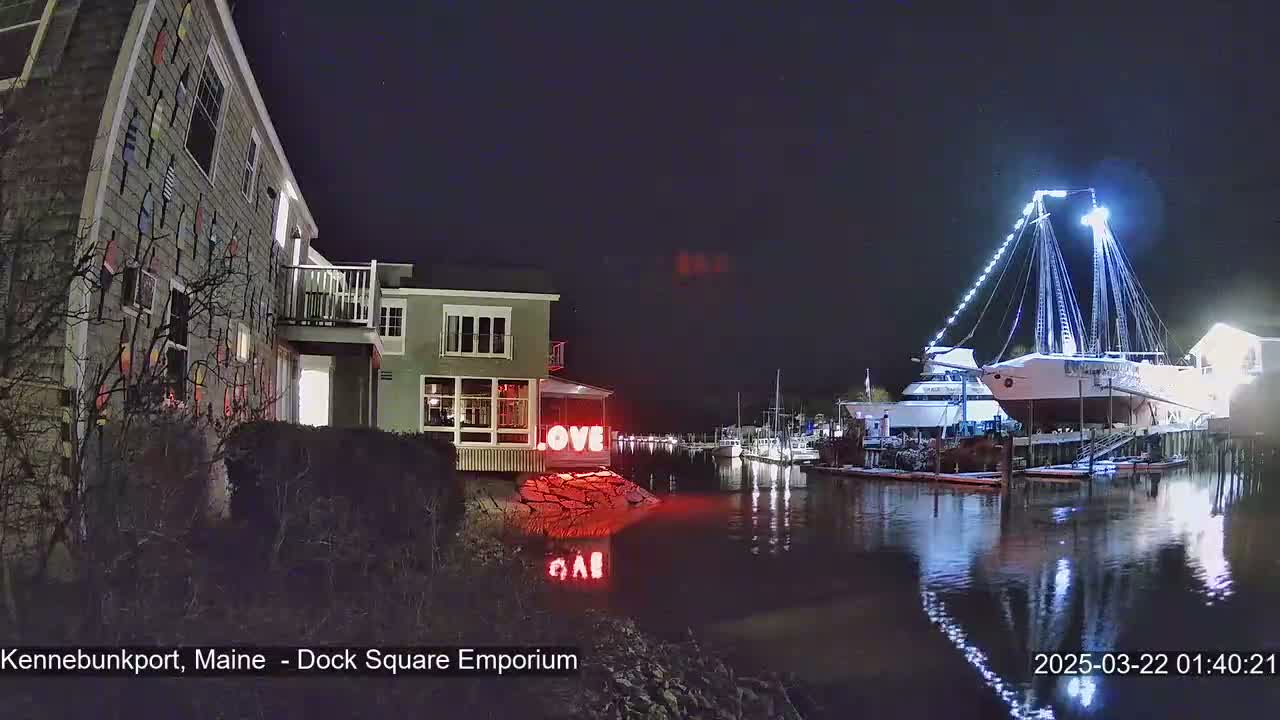 A nighttime waterfront scene shows a building decorated with colorful pennants, another building with illuminated windows, and a large sailing ship docked at a pier, all under a dark, clear sky.