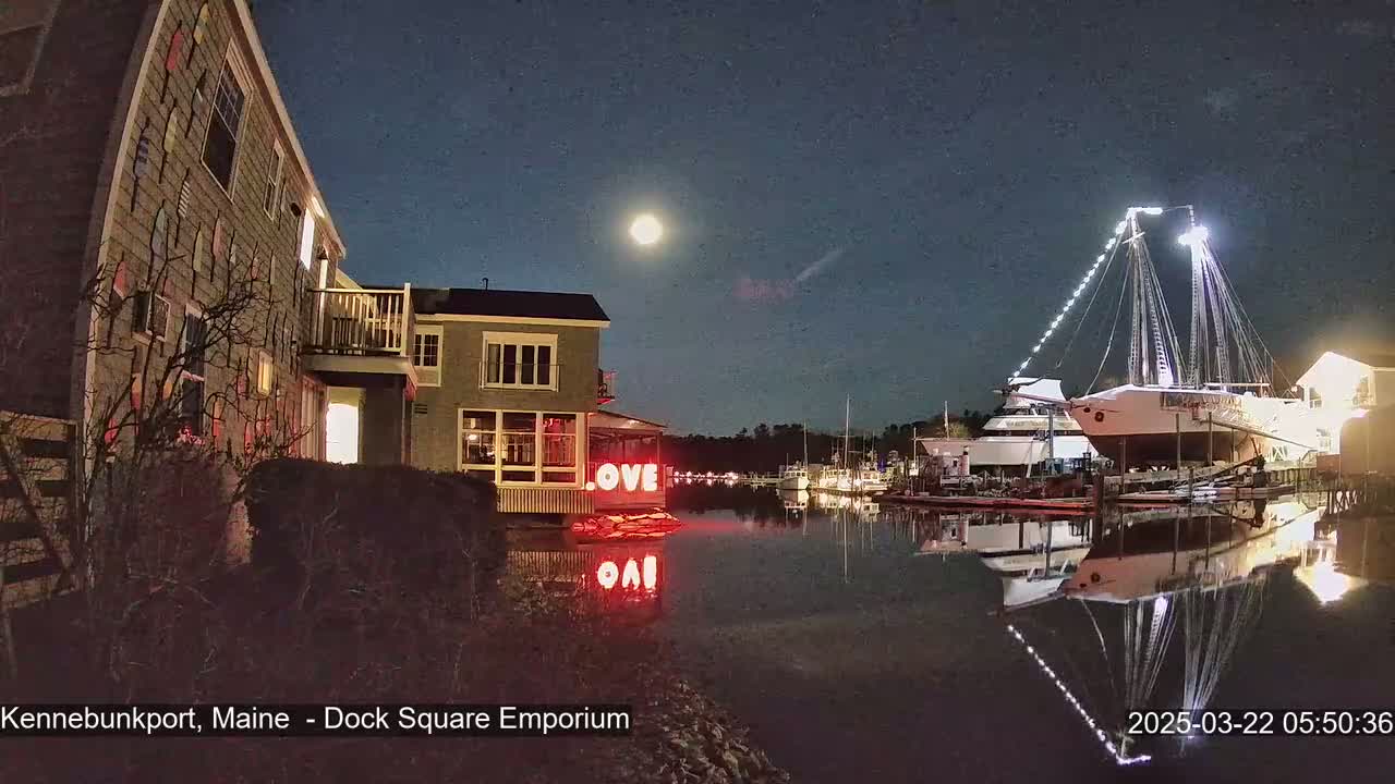 A brightly lit building displaying the word "LOVE" sits on a calm, dark-water harbor at night under a full moon, with a large sailboat illuminated in the background.