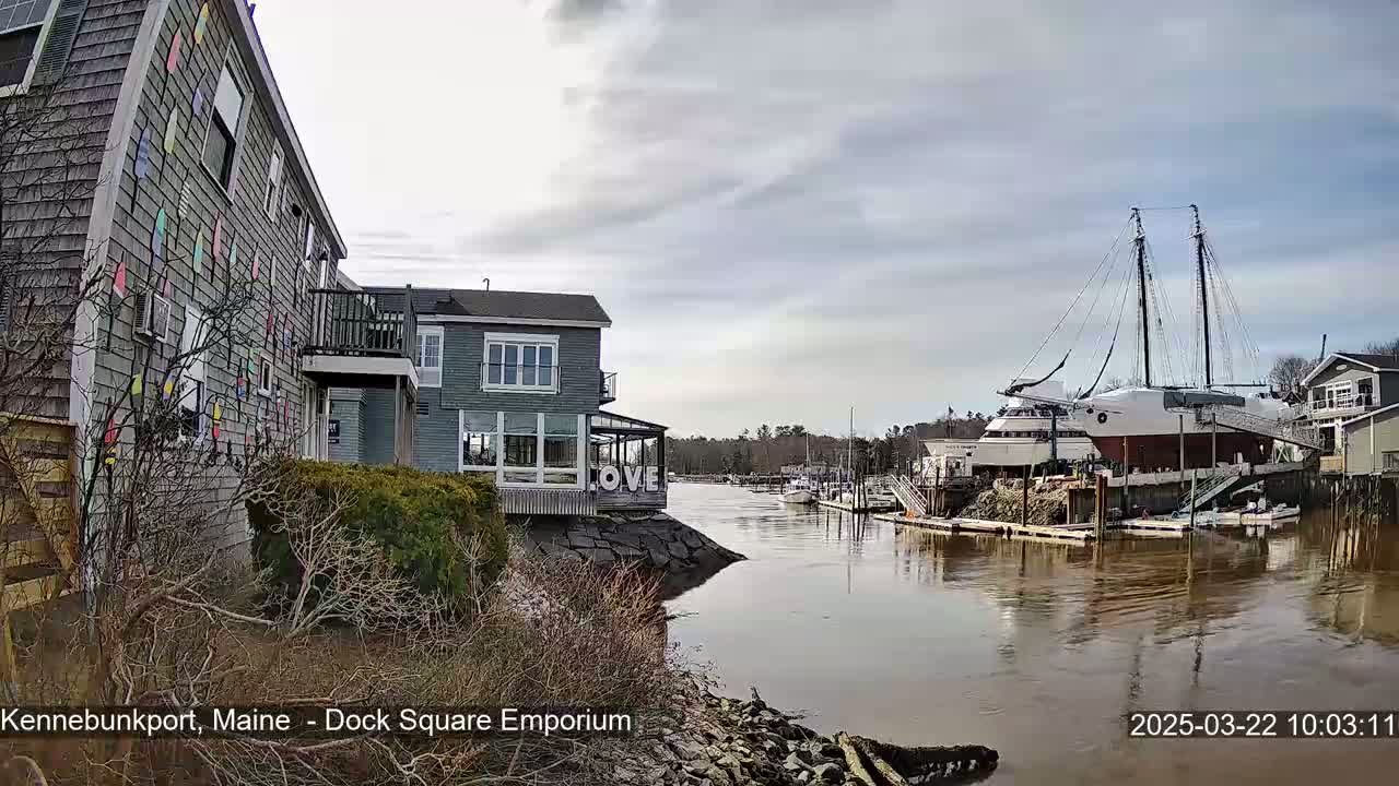 A calm waterway flows between buildings, one adorned with colorful decorations, and a large docked sailing ship under a cloudy sky.