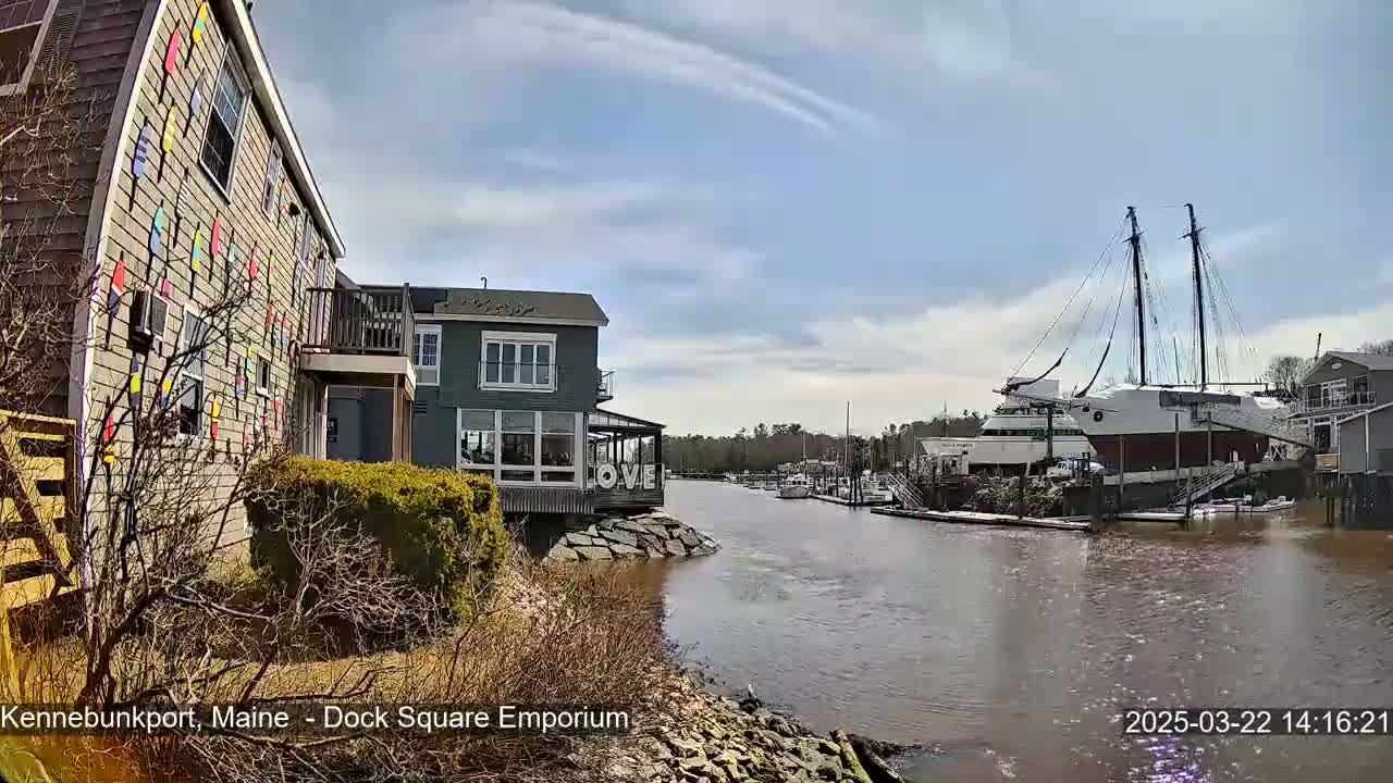 A waterfront scene on a partly cloudy day shows a building decorated with colorful flags next to a calm river where a large sailing ship is docked.