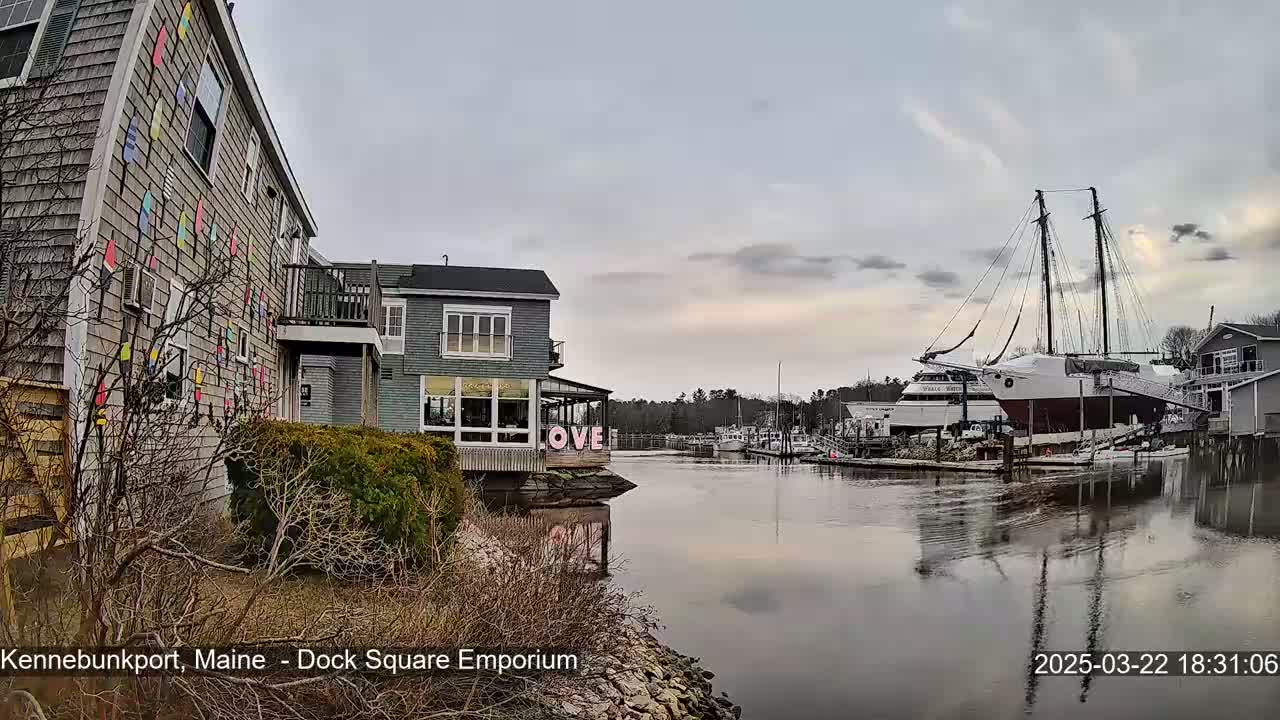 A waterfront scene shows calm water reflecting buildings and a large sailboat, under a cloudy sky.