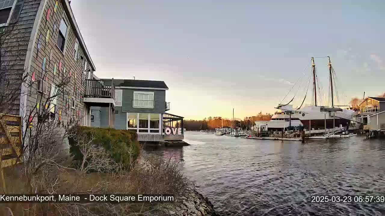 A calm waterway at sunset with a large sailboat docked next to buildings, one of which has colorful painted siding.