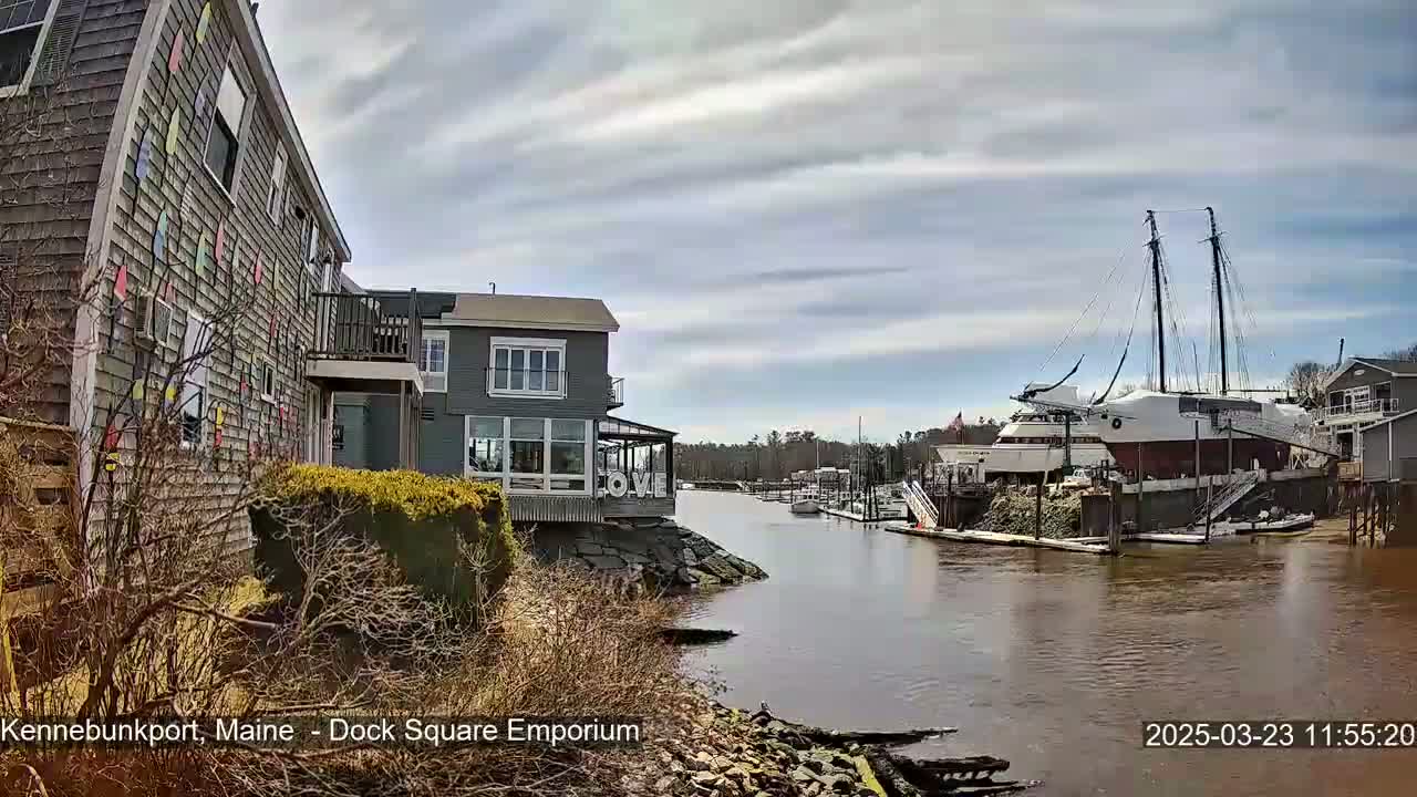 A calm waterway with a large sailing ship docked beside a building decorated with colorful shapes, under a cloudy sky.