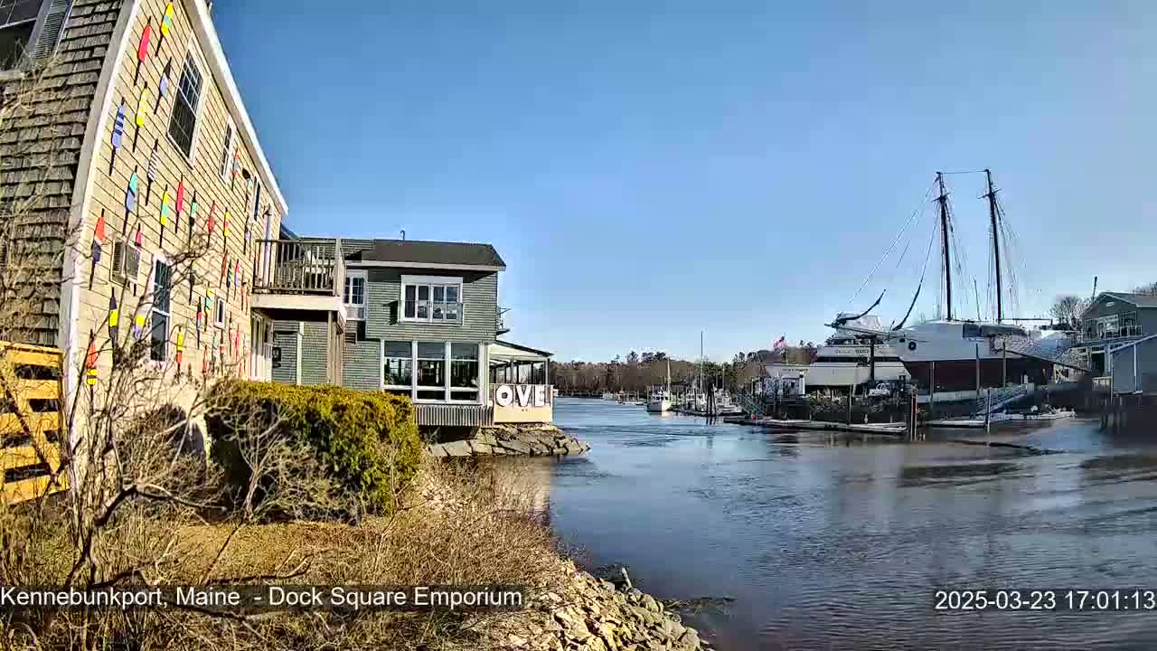 A waterfront scene on a sunny day shows a building decorated with colorful buoys, another building with large windows, and a docked sailing ship, all beside a calm river.