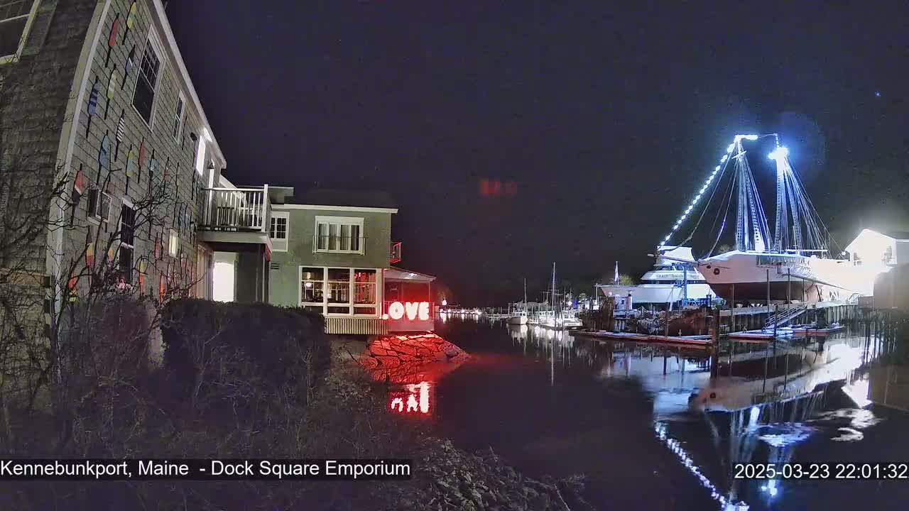 At night, a building with colorful accents stands beside a calm waterway reflecting a docked sailboat illuminated with bright lights, and a red "LOVE" sign is visible on the building's lower level.