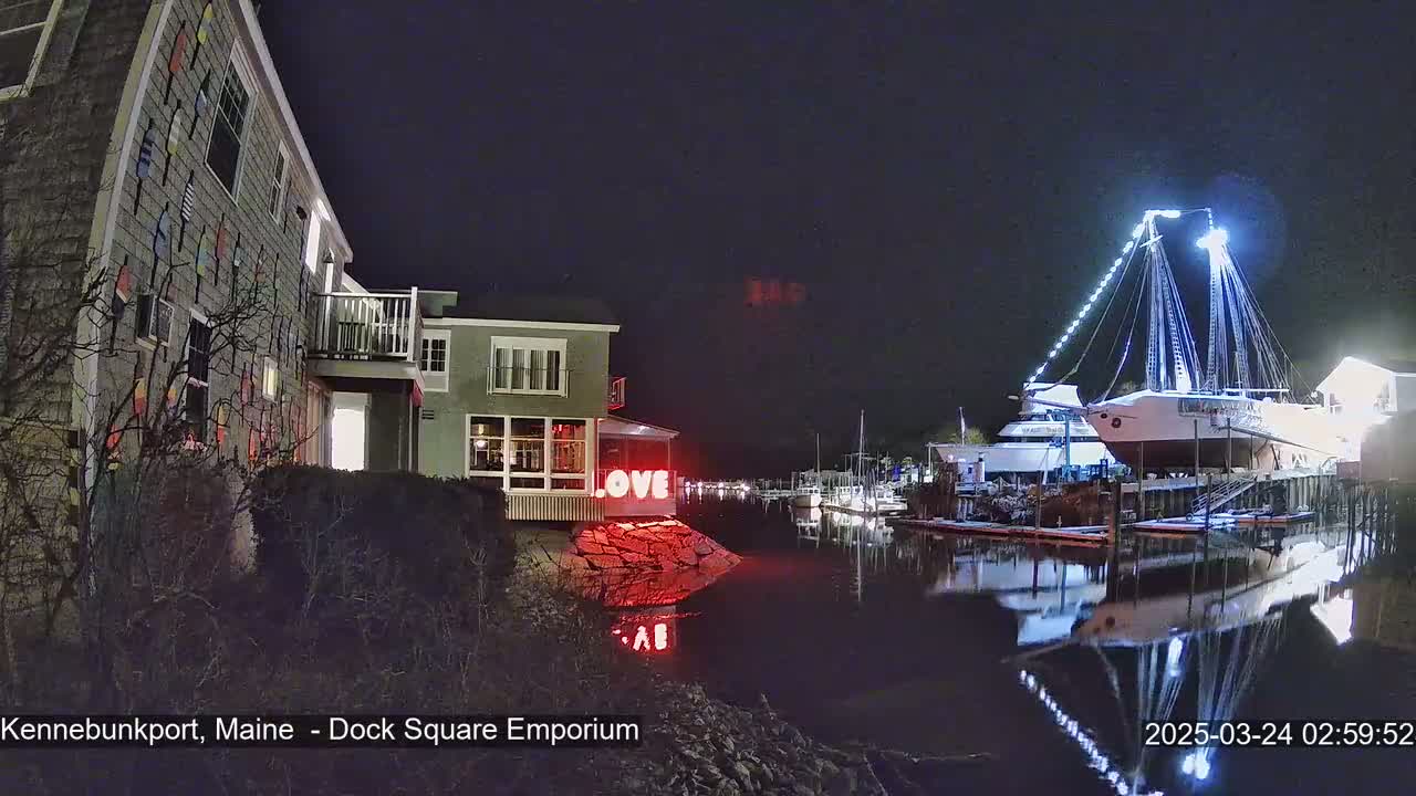 A nighttime view of a waterfront scene with a building decorated with nautical-themed items, a "LOVE" sign, and a large sailboat illuminated at a dock, all reflected in calm water.