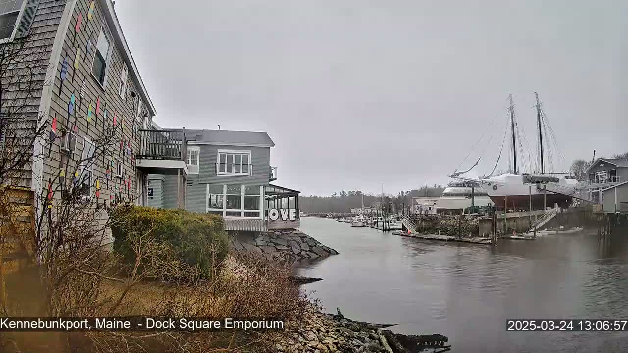 A gray, overcast day reveals a waterfront scene with a building adorned with colorful flags, another building displaying a "LOVE" sign, and a large sailboat docked in a calm harbor.