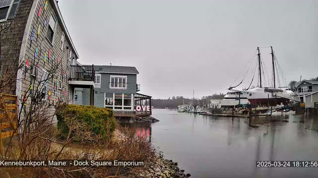 A waterfront view on an overcast day shows a building with colorful nautical flags on its exterior, another building displaying the word "LOVE," and a large sailboat docked in calm water.